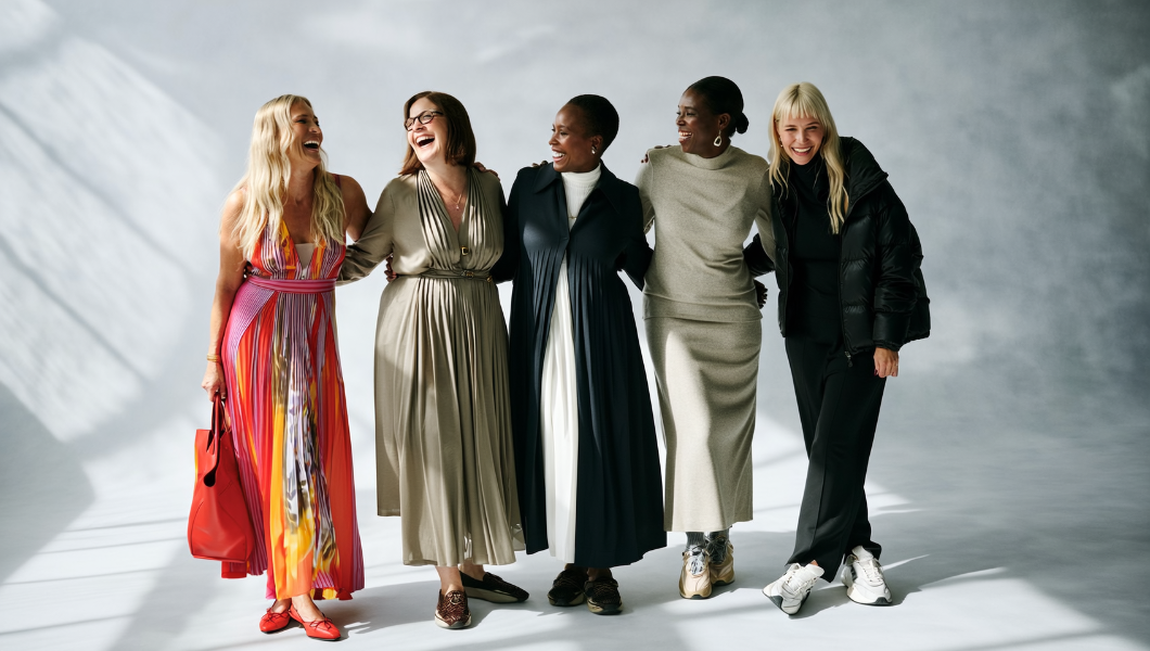 Five women standing together, laughing, against a gray background

